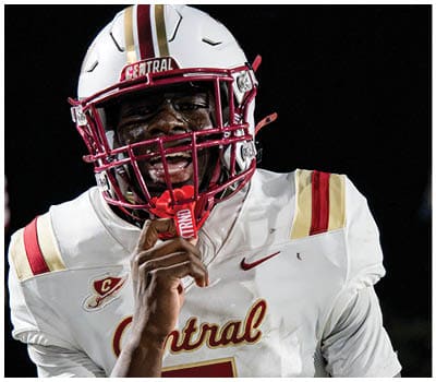 A person wearing a white football helmet with red and gold stripes and a white jersey is smiling with an American flag blurred in the background. Description generated by AI