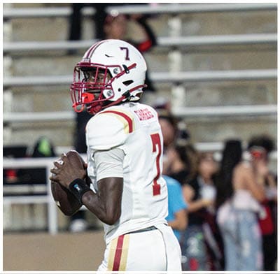 A person wearing a white football uniform with red and gold stripes is holding a football and preparing to throw it on a field with blurred spectators in the background. Description generated by AI