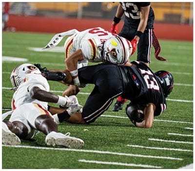 A person wearing a black football uniform and helmet with red and white stripes is on a field at night. Description generated by AI