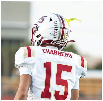A person wearing a white football helmet and jersey with the word Chargers on the back is standing outdoors with a blurred building in the background. Description generated by AI