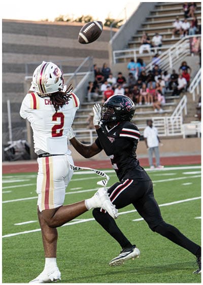 Two people in football uniforms are competing to catch a football in mid-air near stadium bleachers filled with spectators. Description generated by AI