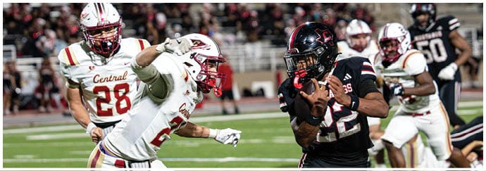 People in football uniforms are actively playing on a field with one person holding the ball while others attempt to block or tackle. Description generated by AI