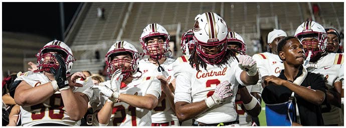 A group of people wearing football helmets and uniforms stand closely together on a field with empty stadium seats in the background. Description generated by AI
