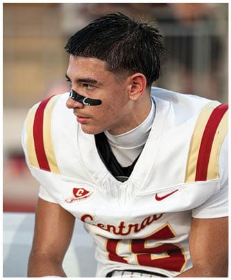 A person wearing a white football jersey with red and gold stripes and black eye paint is sitting and looking to the side. Description generated by AI