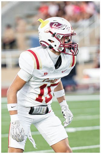 A person wearing a white football helmet and uniform with red and gold accents is focused and ready to play on a blurred stadium background. Description generated by AI