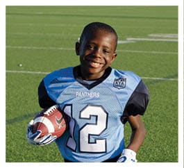 A child wearing a blue and black football uniform with the number 12 is standing on a football field holding a helmet and a football while smiling. Description generated by AI