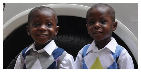 Two children wearing white shirts and argyle sweater vests stand close together in front of a vehicle tire. Description generated by AI