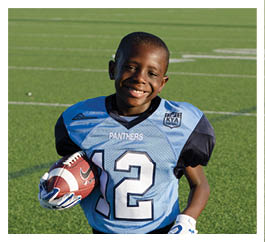 A child wearing a blue and black football uniform with the number 12 is standing on a football field holding a helmet and a football while smiling. Description generated by AI
