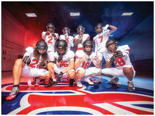 Four people wearing football helmets and uniforms crouch on a gym floor with a large colorful logo, posing with hand gestures. Description generated by AI