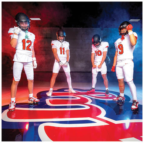 Four people wearing football uniforms and helmets stand on a gym floor with colored smoke in the background, posing confidently around a large logo. Description generated by AI