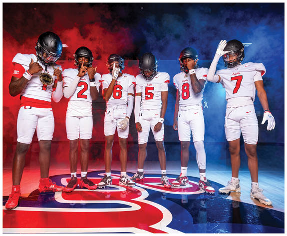 Six people wearing white football uniforms with black helmets stand on a colorful floor with red and blue smoke in the background, each posing with one hand near their face or raised. Description generated by AI