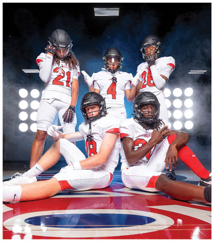 Five people in white football uniforms and helmets pose confidently on a lit indoor field with a blue and red floor design. Description generated by AI