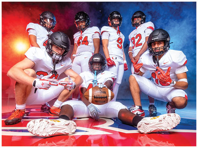 Seven people in football uniforms and helmets pose confidently with one holding a football in a dramatic setting with red and blue lighting. Description generated by AI