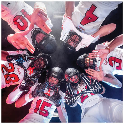 A group of six people wearing football helmets and uniforms are huddled in a circle looking down at the camera with some making hand gestures. Description generated by AI