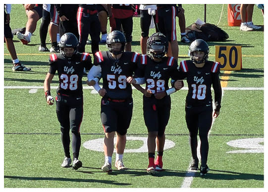 Two people wearing black football uniforms with the word “Ugly" on the front are standing side by side on a field with their arms linked. Description generated by AI