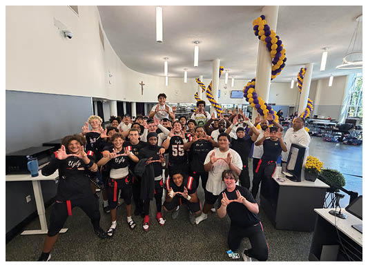 A group of people wearing sports attire are gathered indoors, smiling and making a hand gesture that forms a “U" shape. Description generated by AI