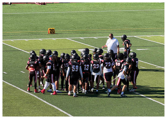 A group of people wearing black football uniforms with helmets are gathered on a green field while a person in a white shirt stands nearby. Description generated by AI