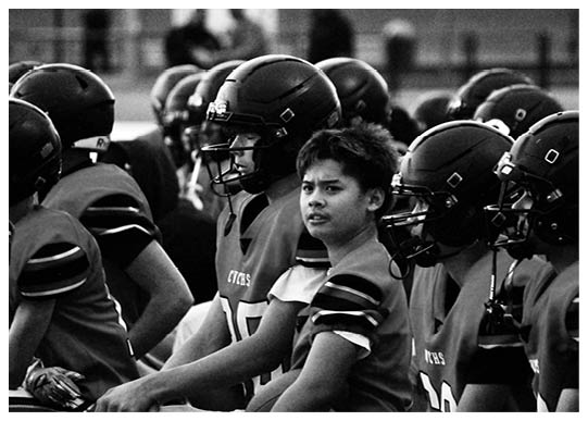 A group of people wearing football helmets and jerseys are sitting closely together, with one person looking directly at the camera. Description generated by AI