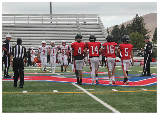Four people in red football uniforms walk arm in arm on a football field toward a group of people in white uniforms while referees stand nearby. Description generated by AI