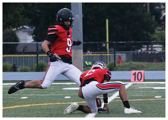 Two people in red and white football uniforms are on a field with one person holding the ball on the ground while the other is kicking it. Description generated by AI