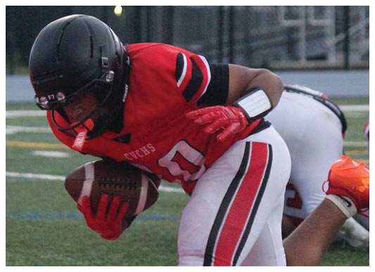A person wearing a red and white football uniform with a black helmet is holding a football and bending forward on a grassy field. Description generated by AI