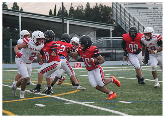 Several people wearing football uniforms are actively engaged in a game on a green field with yard lines and a scoreboard visible in the background. Description generated by AI