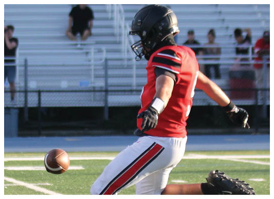 A person wearing a red and white football uniform with a black helmet is holding a football and bending forward on a grassy field. Description generated by AI