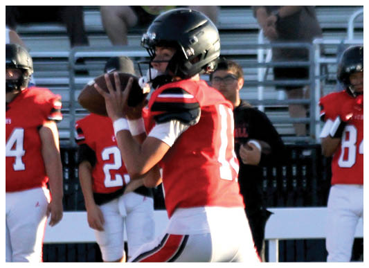 A person in a red and white football uniform and black helmet is holding a football in a throwing position with other players and a person in casual clothing in the background. Description generated by AI