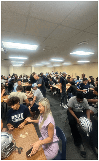 A large group of people are seated and standing around tables in a brightly lit room, engaging in conversation and activities. Descripci n generada con IA