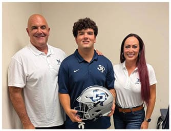 Three people wearing polo shirts with a sports logo pose indoors, with the person in the center holding a football helmet. Descripci n generada con IA
