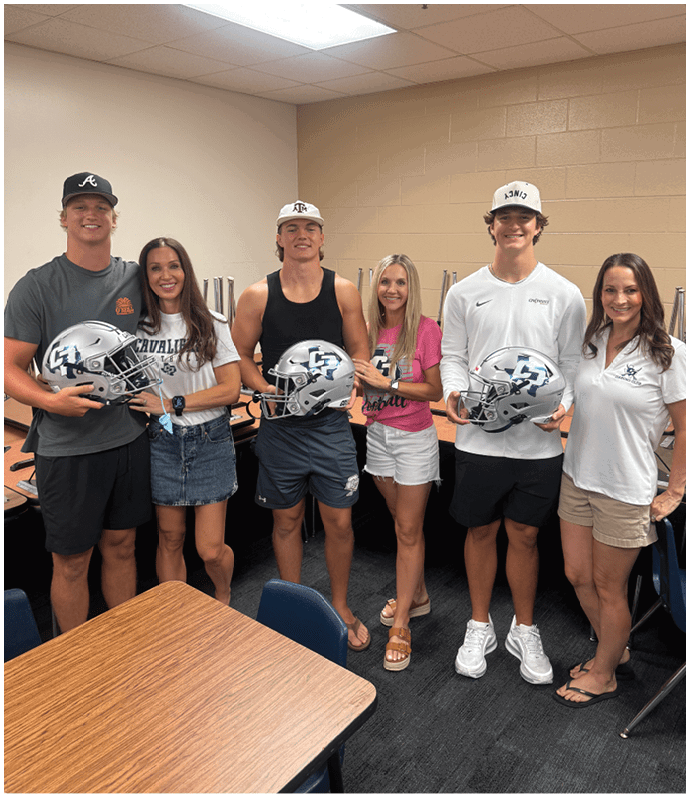 Four people standing indoors smiling and holding football helmets, with two wearing casual t-shirts and two wearing athletic clothing and caps. Descripci n generada con IA