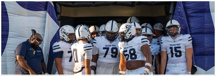 A group of people wearing white football uniforms with helmets stand together near an inflatable tunnel while a person in a dark shirt looks down nearby. Descripci n generada con IA
