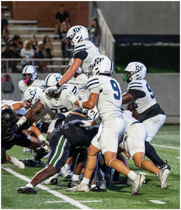 A group of people in white and dark football uniforms are engaged in a play on a green field with spectators in the background. Descripci n generada con IA