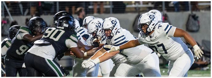 Two opposing American football teams in helmets and uniforms are lined up facing each other on the field, preparing for a play. Descripci n generada con IA