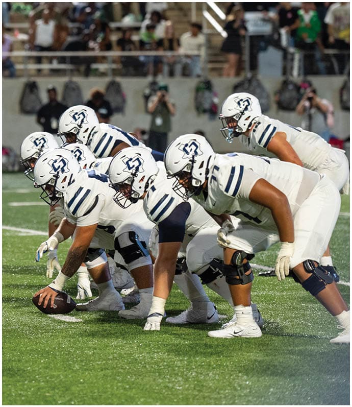 A group of football players in white uniforms are lined up on the field facing a player in a black helmet and dark uniform, preparing for a play. Descripci n generada con IA