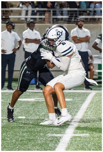 Two people in football uniforms are engaged in a tackle on a football field with coaches and players watching in the background. Descripci n generada con IA