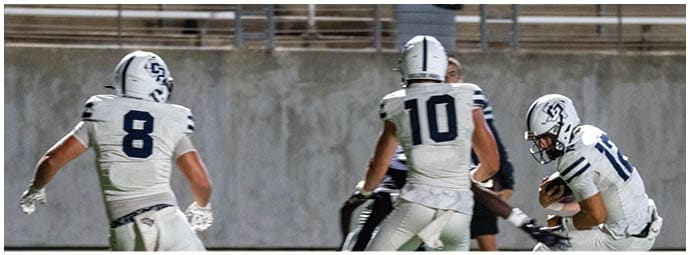 Three people in football uniforms are on a field with one person holding a football while the others appear to be running or preparing to block. Descripci n generada con IA