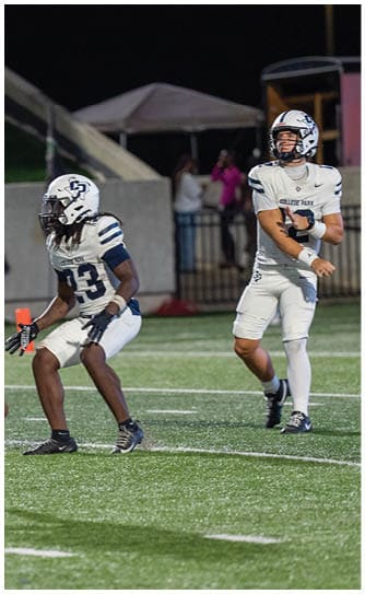 Two people in white football uniforms with helmets are on a field at night, with one preparing to throw a football and the other in a ready stance. Descripci n generada con IA
