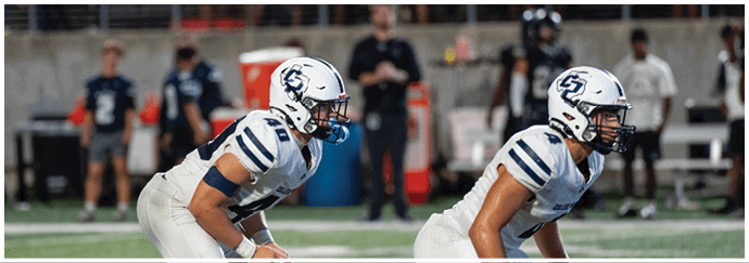 Two people in football uniforms and helmets are crouched in a ready position on a football field during a game. Descripci n generada con IA