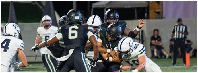 People wearing football helmets and uniforms are engaged in a play on a football field at night with a referee and spectators in the background. Descripci n generada con IA