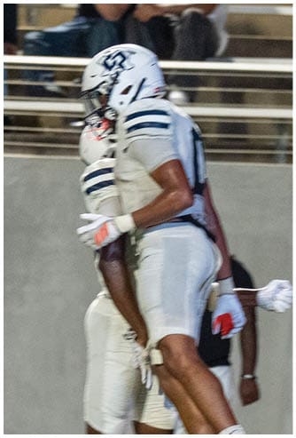 Two people wearing white football uniforms and helmets are standing close together on a field with spectators in the background. Descripci n generada con IA