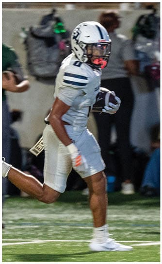 A person in a white football uniform is running on a field while holding a football, with other players and spectators visible in the background. Descripci n generada con IA