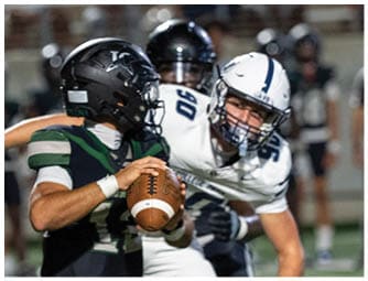 A person in a dark sports uniform is holding a football while another person in a white sports uniform is reaching toward them on a grassy field during a game. Descripci n generada con IA