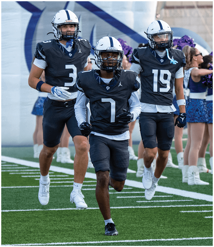 Three people in football uniforms are running on a field with cheerleaders in blue and white outfits standing in the background. Descripci n generada con IA