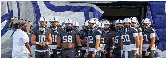 A group of people wearing matching dark sports uniforms and helmets stand together on a field, appearing to prepare for a game. Descripci n generada con IA