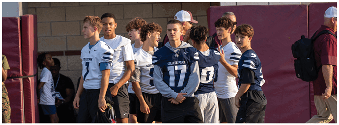 A group of people wearing sports jerseys and shorts are standing and talking near a sports field with a brick wall and maroon padding in the background. Descripci n generada con IA