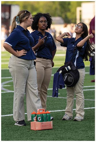 Three adults wearing navy blue polo shirts and khaki pants stand on a sports field with water bottles and a backpack nearby, engaged in conversation. Descripci n generada con IA