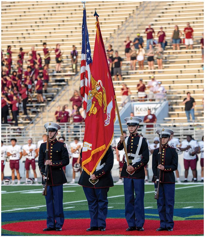 Two flags, one American and one Marine Corps, are held upright in front of a stadium with people standing in the bleachers. Descripci n generada con IA