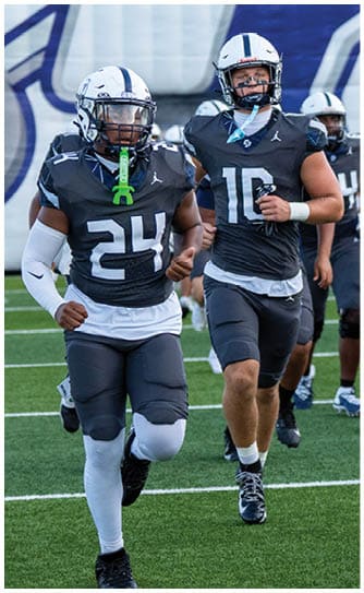Two people wearing dark football uniforms with white helmets are running on a field during a game. Descripci n generada con IA