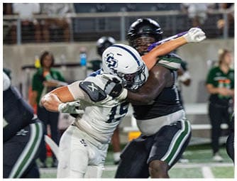 Two football players in contrasting uniforms are engaged in a physical play on a field with other players and spectators in the background. Descripci n generada con IA